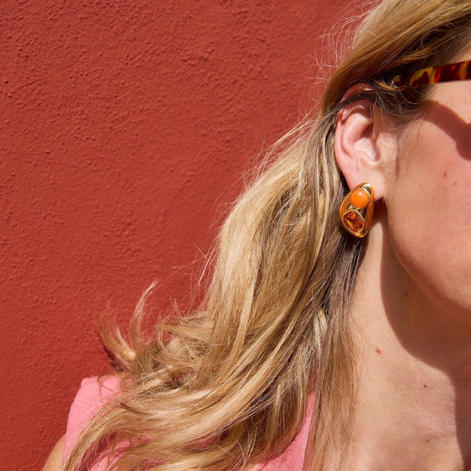 Close-up of a person wearing a gold earring with an orange gemstone against a red wall.