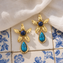 A pair of gold dangle earrings with textured gold petals, deep blue round stones at the center, and teardrop-shaped turquoise stones hanging below, displayed on blue-and-white patterned tiles with a white fabric drape.