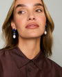 A close-up of a woman wearing blue and white drop earrings with a dark brown button-down shirt, set against a plain light gray background.