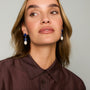 A close-up of a woman wearing blue and white drop earrings with a dark brown button-down shirt, set against a plain light gray background.