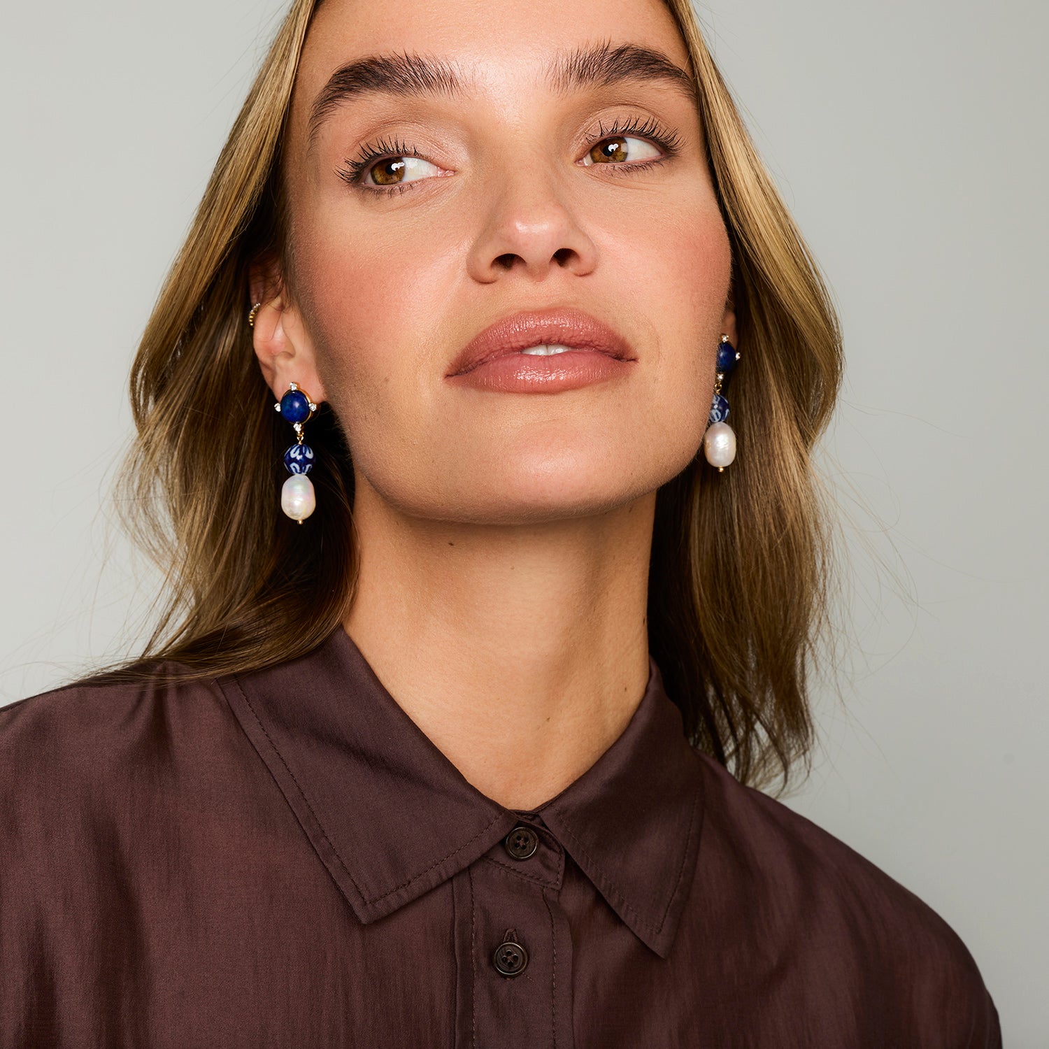A close-up of a woman wearing blue and white drop earrings with a dark brown button-down shirt, set against a plain light gray background.