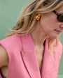 Woman wearing a pink sleeveless blazer with sunglasses and gold and glass stone earrings against a green background