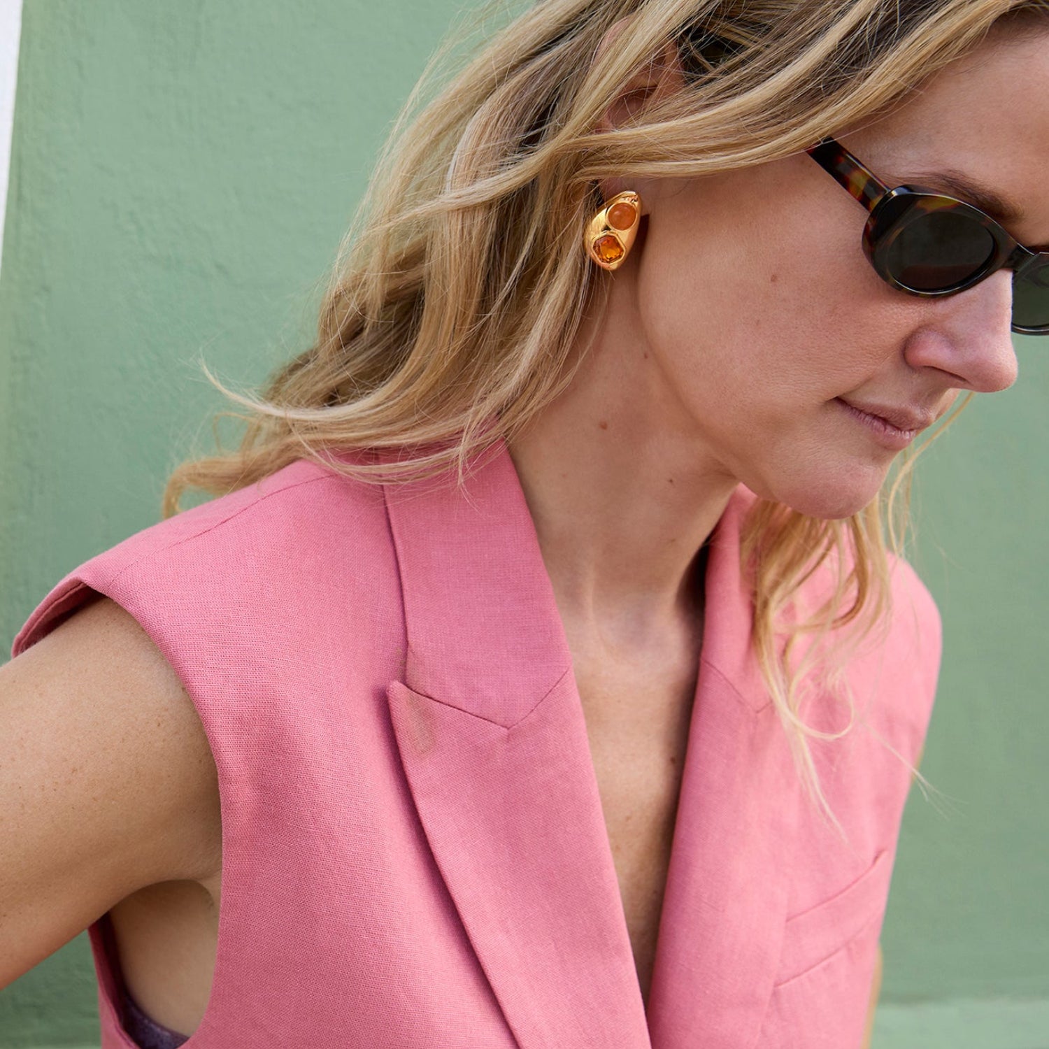 Woman wearing a pink sleeveless blazer with sunglasses and gold and glass stone earrings against a green background
