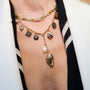 A woman wearing a white top, and a gold-toned link necklace with a pearl and amber pendant, paired with a green and amber colored tennis necklace