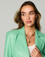 A woman with light brown hair wearing a mint green leather blazer, white top, and a gold-toned beaded necklace with a pearl and amber pendant, photographed against a solid light gray background.