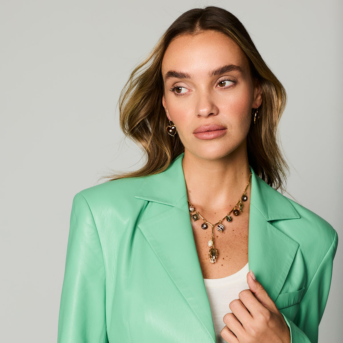 A woman with light brown hair wearing a mint green leather blazer, white top, and a gold-toned beaded necklace with a pearl and amber pendant, photographed against a solid light gray background.