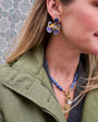 A close-up lifestyle shot of a woman wearing a pair of colorful butterfly-shaped earrings with blue, purple, and yellow enamel details, along with a blue beaded necklace and an olive green jacket.