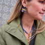 A close-up lifestyle shot of a woman wearing a pair of colorful butterfly-shaped earrings with blue, purple, and yellow enamel details, along with a blue beaded necklace and an olive green jacket.