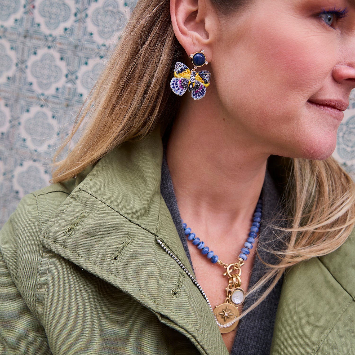 A close-up lifestyle shot of a woman wearing a pair of colorful butterfly-shaped earrings with blue, purple, and yellow enamel details, along with a blue beaded necklace and an olive green jacket.