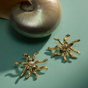 Brushed Wavy Gold and Crystal Sun Star Earrings Staged on Green Surface with a Shell