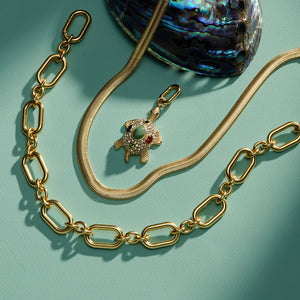 Crystal and Stone Turtle Charm Staged with Herringbone Gold Necklace and Gold Chain Necklace with Shell on Green Surface
