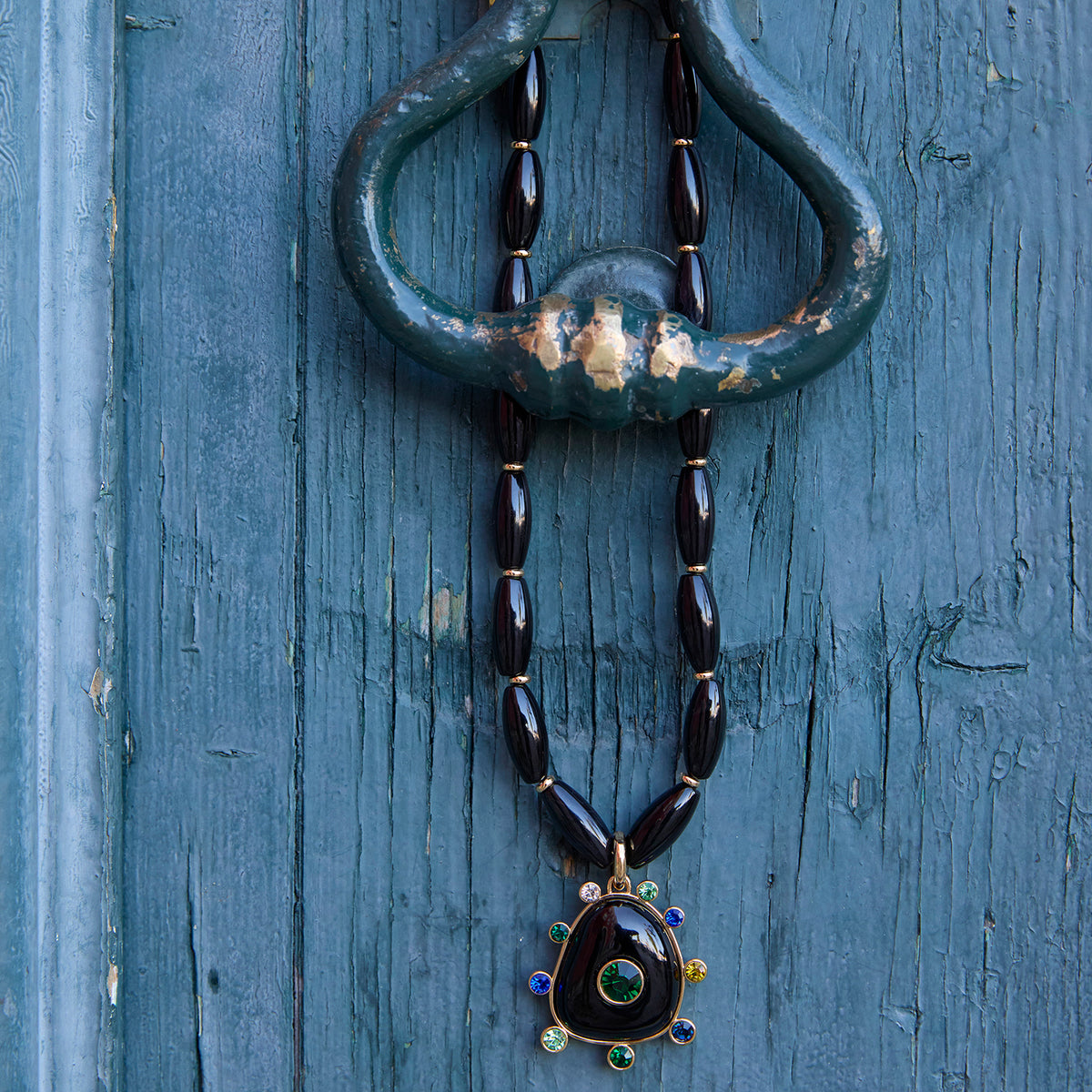 Black beaded necklace featuring a gold-tone Semi Precious Black Onyx pendant with green, navy, blue and clear accents Lay Down Shot on a blue background