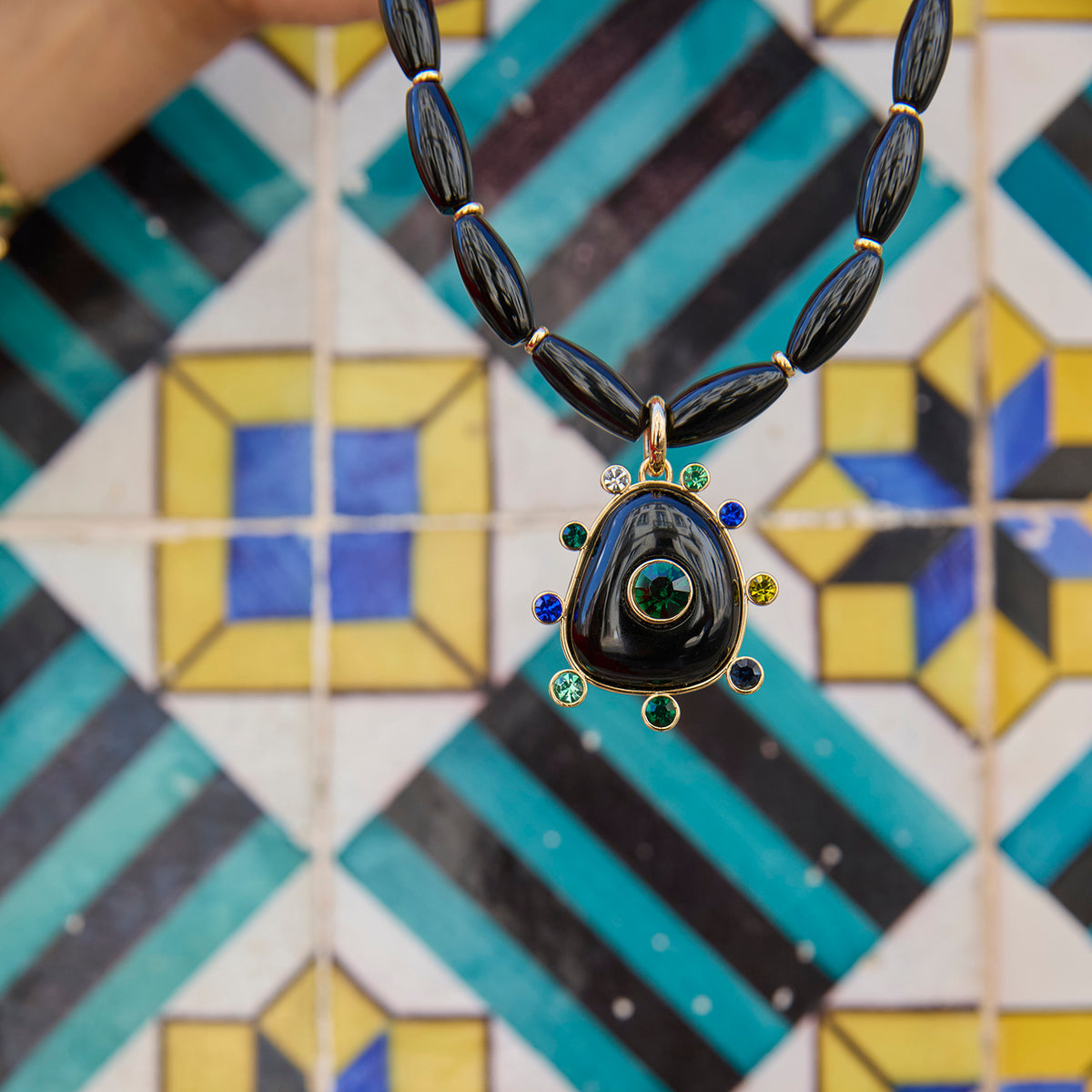 Black beaded necklace featuring a gold-tone Semi Precious Black Onyx pendant with green, navy, blue and clear accents on tile background
