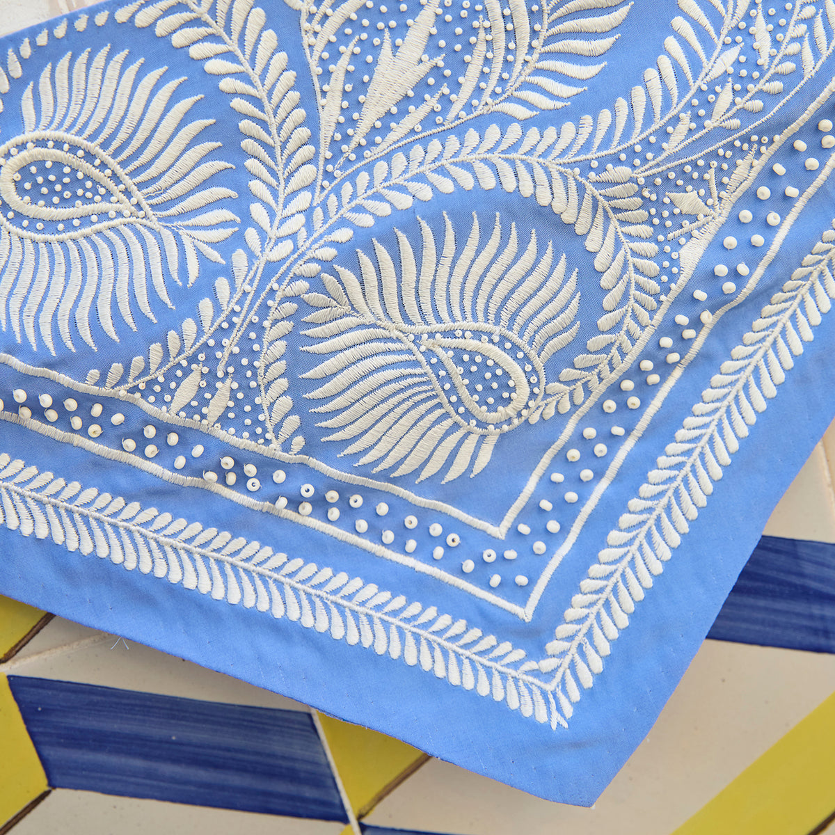 A close-up of a light blue fabric bandana or napkin featuring white block-printed paisley and leaf patterns, laid diagonally over a tiled surface.