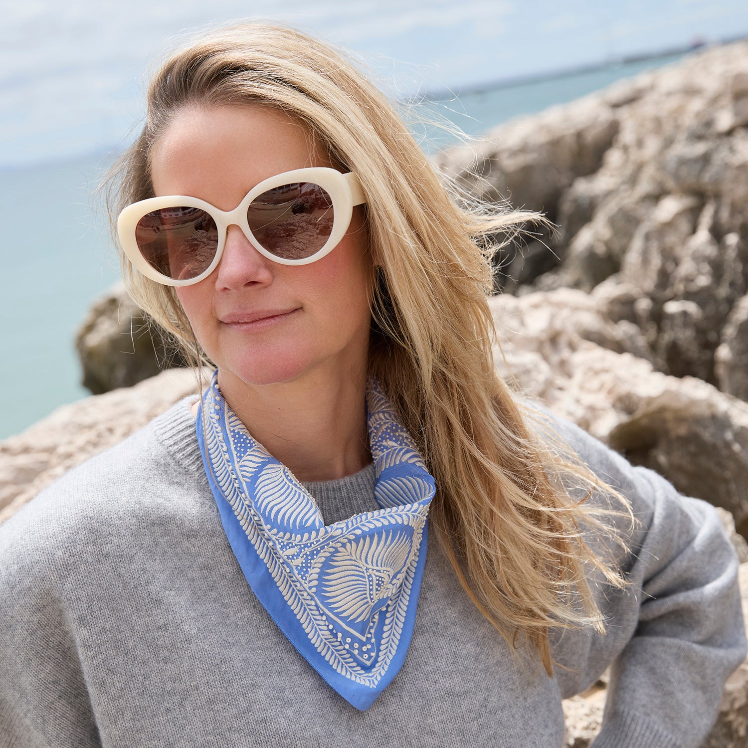 A woman outdoors on a rocky coast wearing a light blue and white patterned bandana neck scarf, a gray sweater, and cream-colored sunglasses.