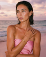 Woman in a pink strapless top and gold cuff bracelet on a beach with a sunset in the background