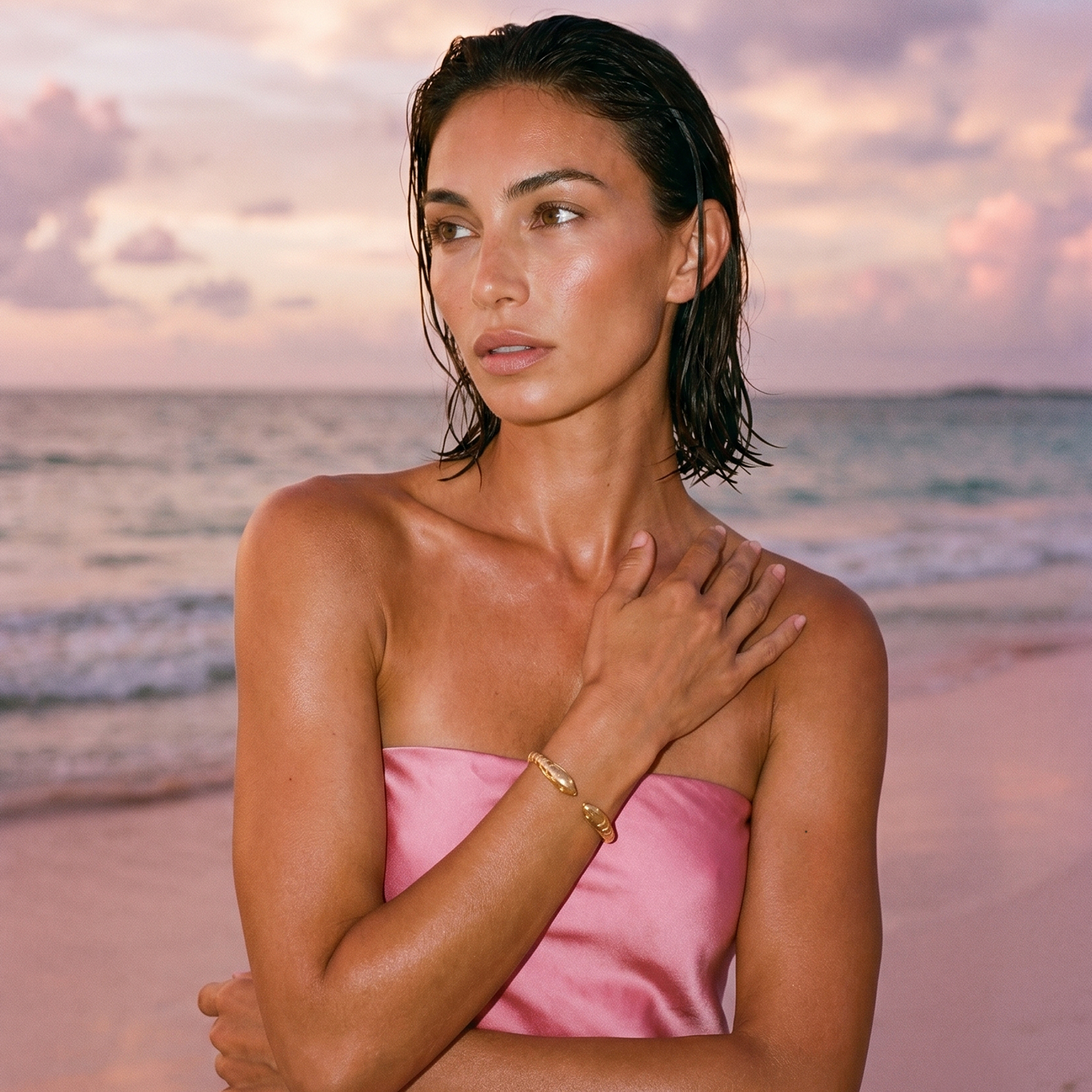 Woman in a pink strapless top and gold cuff bracelet on a beach with a sunset in the background
