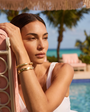 Woman relaxing on a lounge chair by a pool with palm trees in the background with gold cuff bracelets on wrist