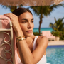 Woman relaxing on a lounge chair by a pool with palm trees in the background with gold cuff bracelets on wrist