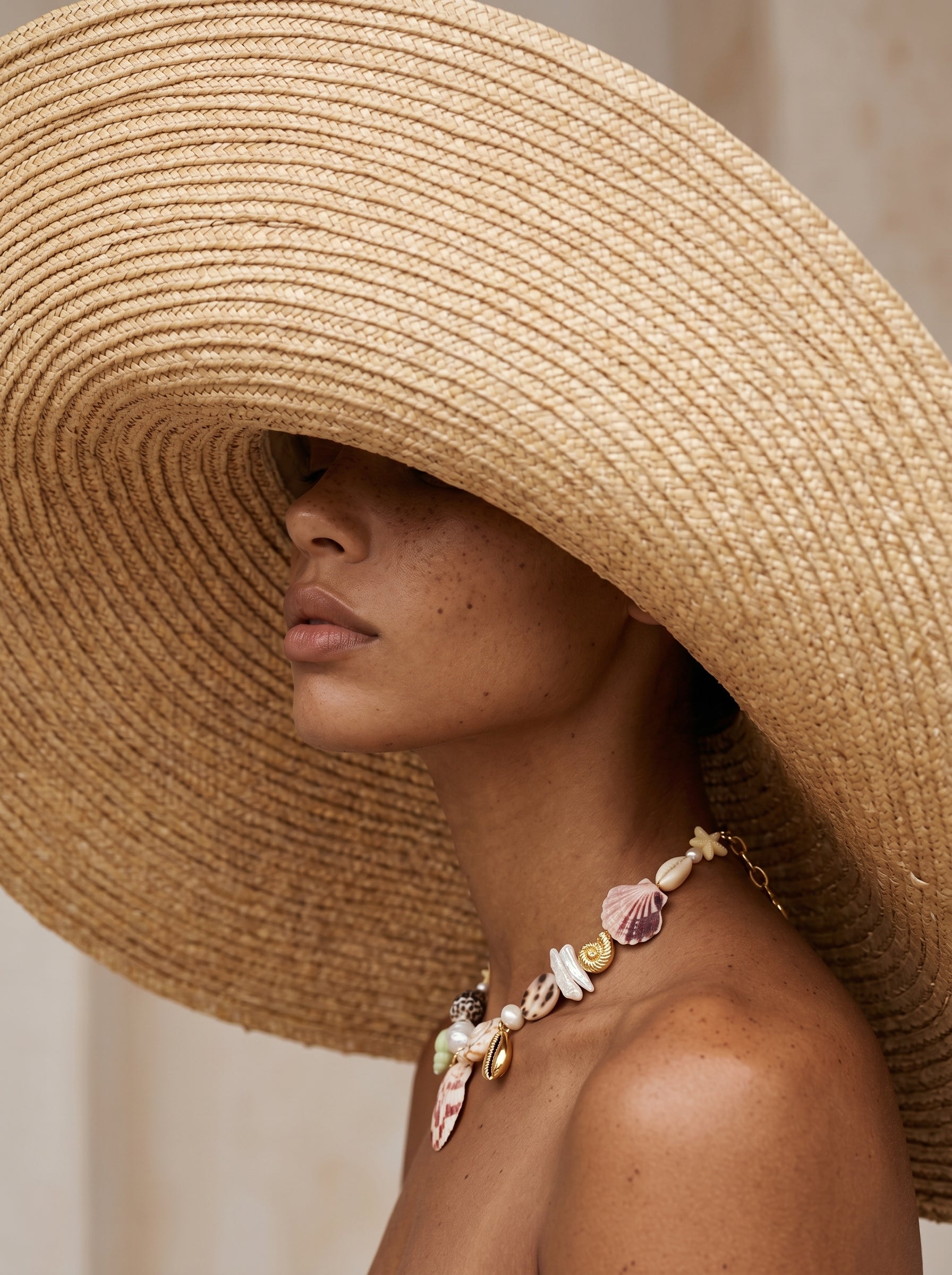 Model wearing large raffia sun hat covering face with large shell charm necklace around her neck