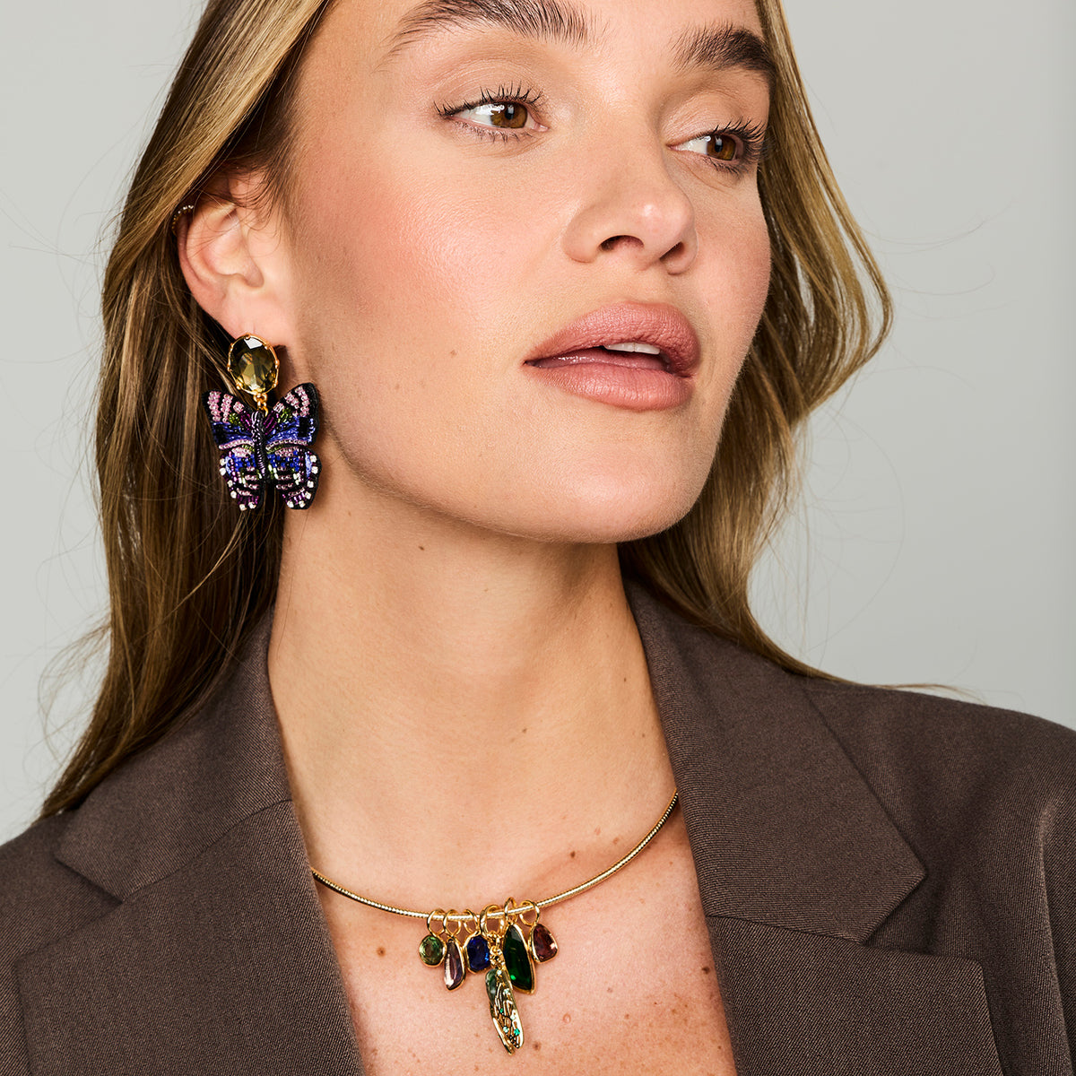 Multicolor butterfly-shaped statement earrings featuring purple, blue, and green beadwork with gold-tone details on Model looking left