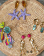 Assortment of statement earrings on a round woven raffia mat against a gray stone background