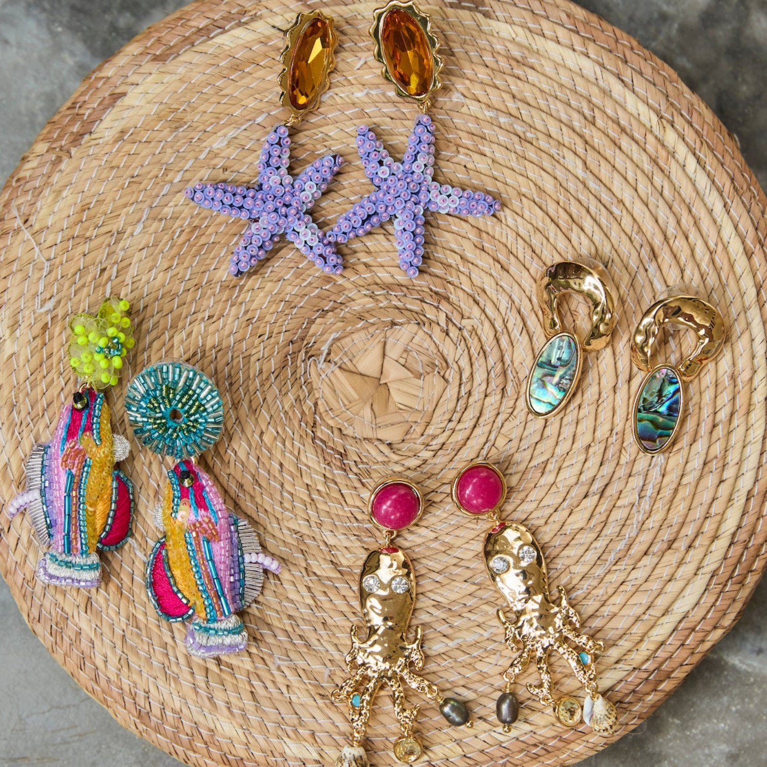 Assortment of statement earrings on a round woven raffia mat against a gray stone background
