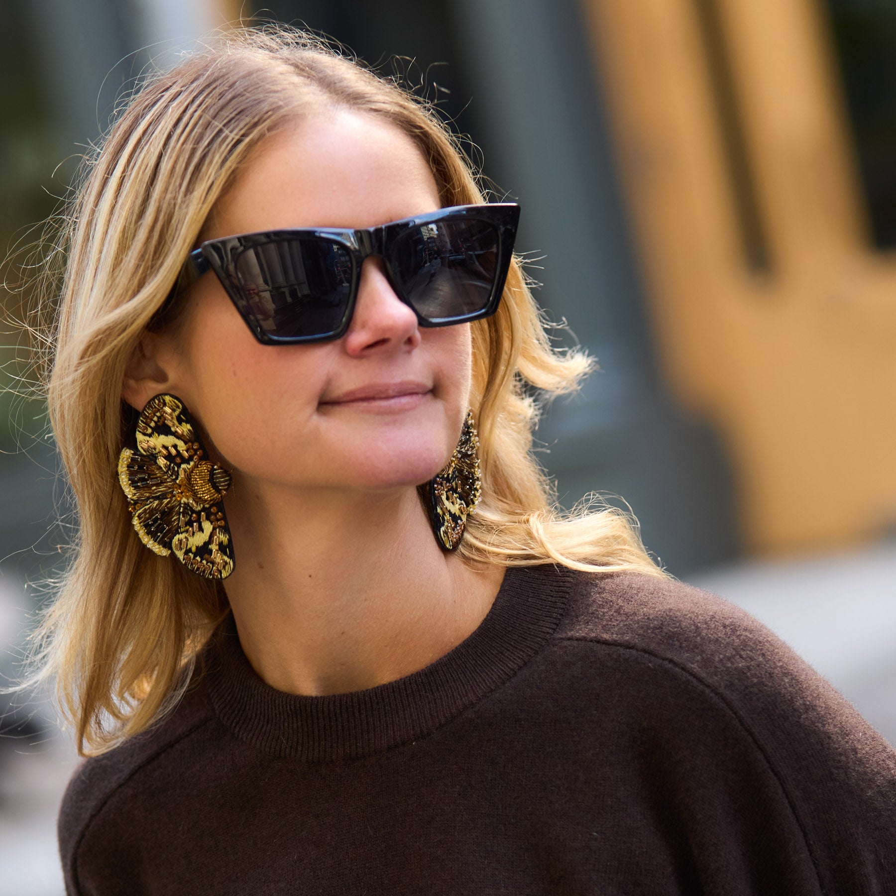 Maggie wearing sunglasses, large moth earrings, and a dark sweater with a blurred background