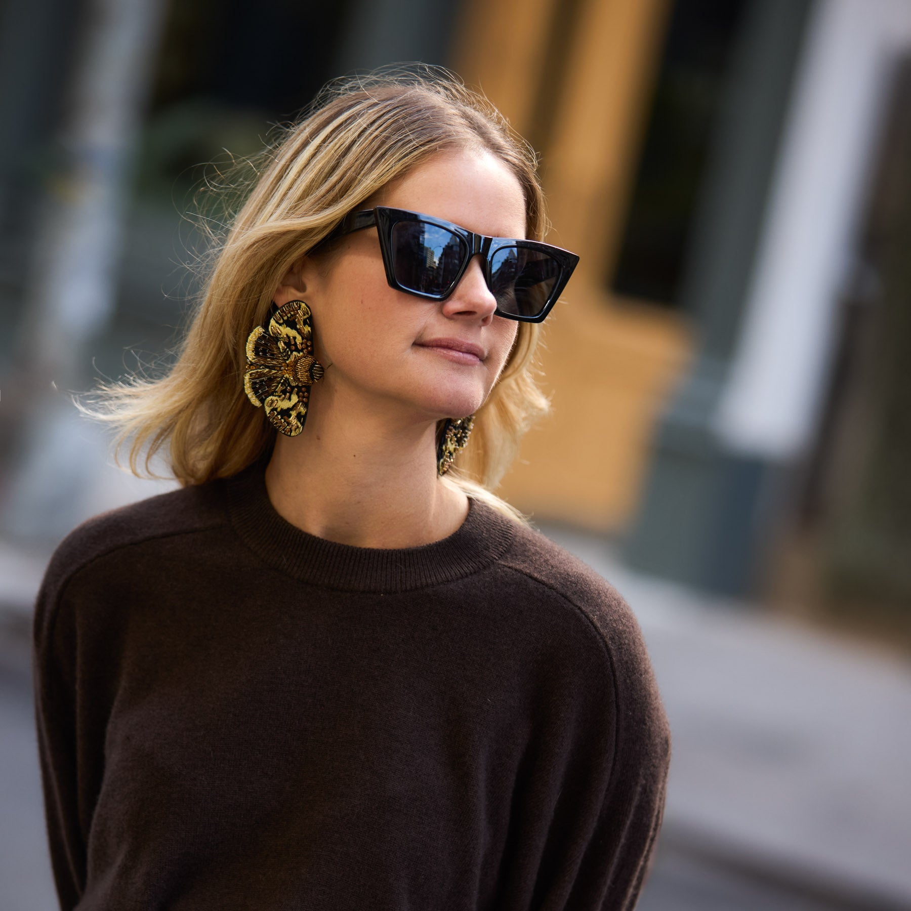 Maggie wearing sunglasses, large moth earrings, and a dark sweater with a blurred background