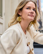 Woman wearing a cream-colored blazer with gold buttons and earrings, standing outdoors.