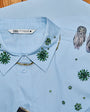 A light blue button-up shirt by Zara, featuring green and gold embroidered floral patterns and a decorative gold chain detail along the collar. A pair of owl-shaped feather earrings is draped on the right side of the shirt, which is laid flat on a wooden surface.