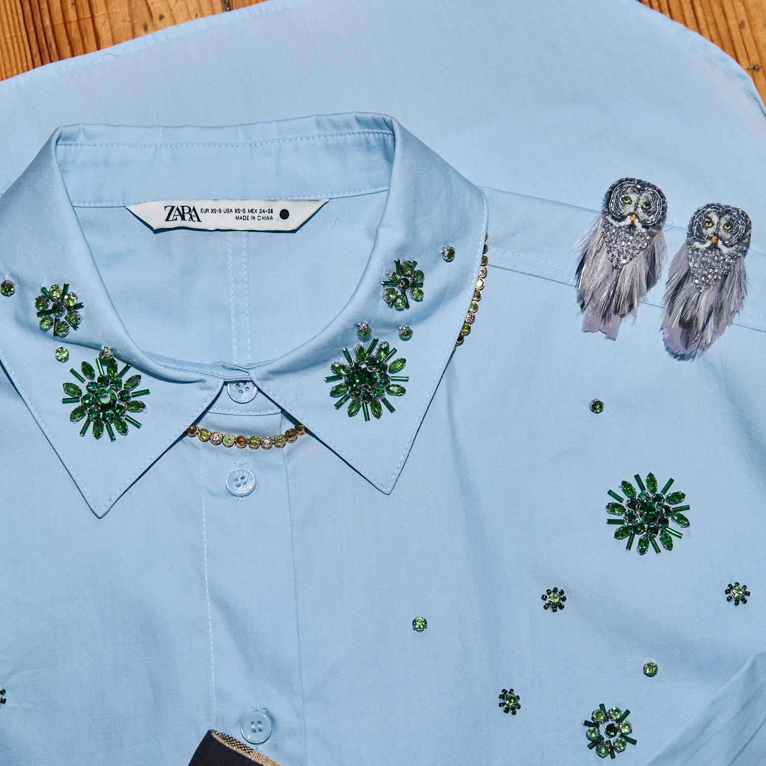 A light blue button-up shirt by Zara, featuring green and gold embroidered floral patterns and a decorative gold chain detail along the collar. A pair of owl-shaped feather earrings is draped on the right side of the shirt, which is laid flat on a wooden surface.