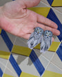 A pair of owl-shaped earrings with gray and white feathers, held in a person's hand against a blue and yellow geometric tiled background.