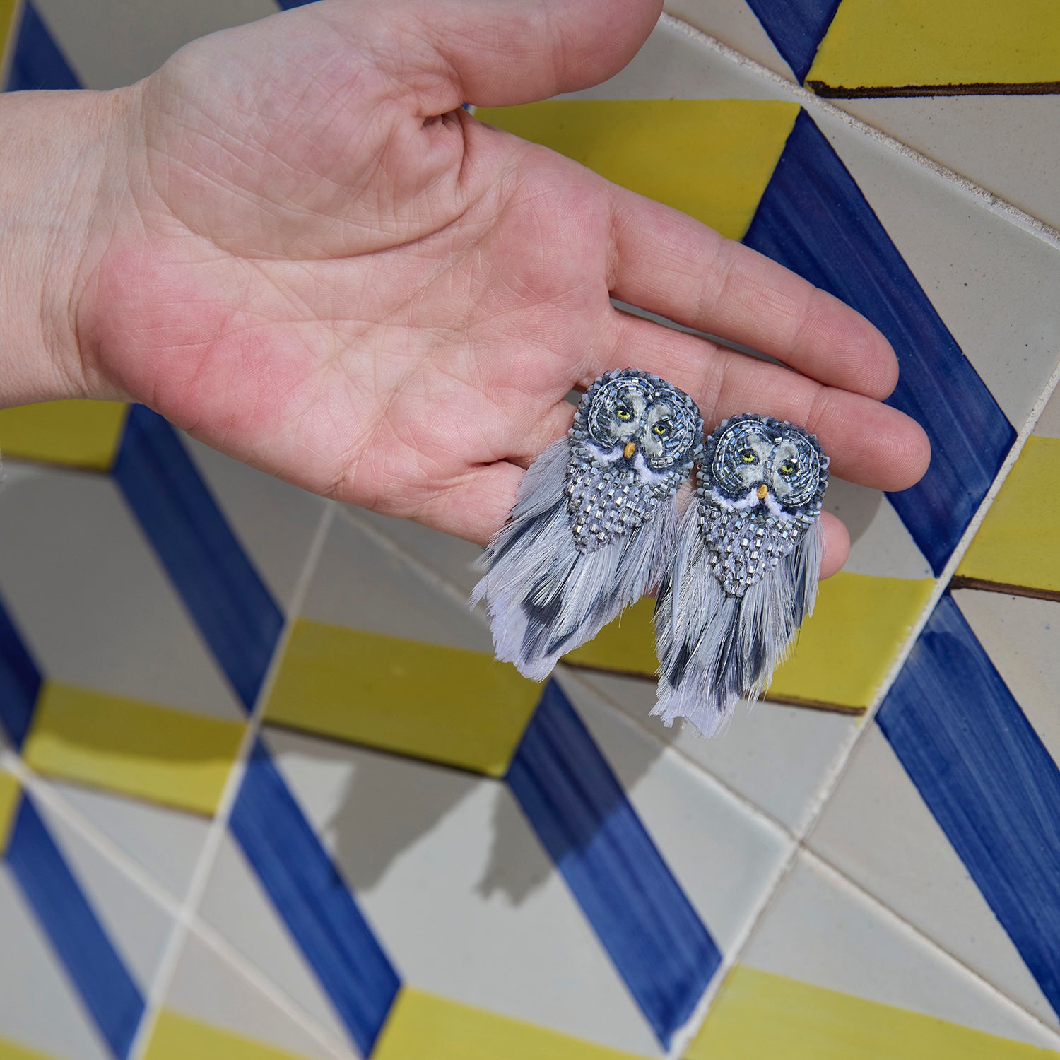 A pair of owl-shaped earrings with gray and white feathers, held in a person's hand against a blue and yellow geometric tiled background.
