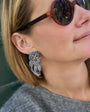 A close-up lifestyle shot of a woman wearing a silver owl-shaped dangle earring with white feathers, shown on her ear. She is wearing round sunglasses and a gray knit top.