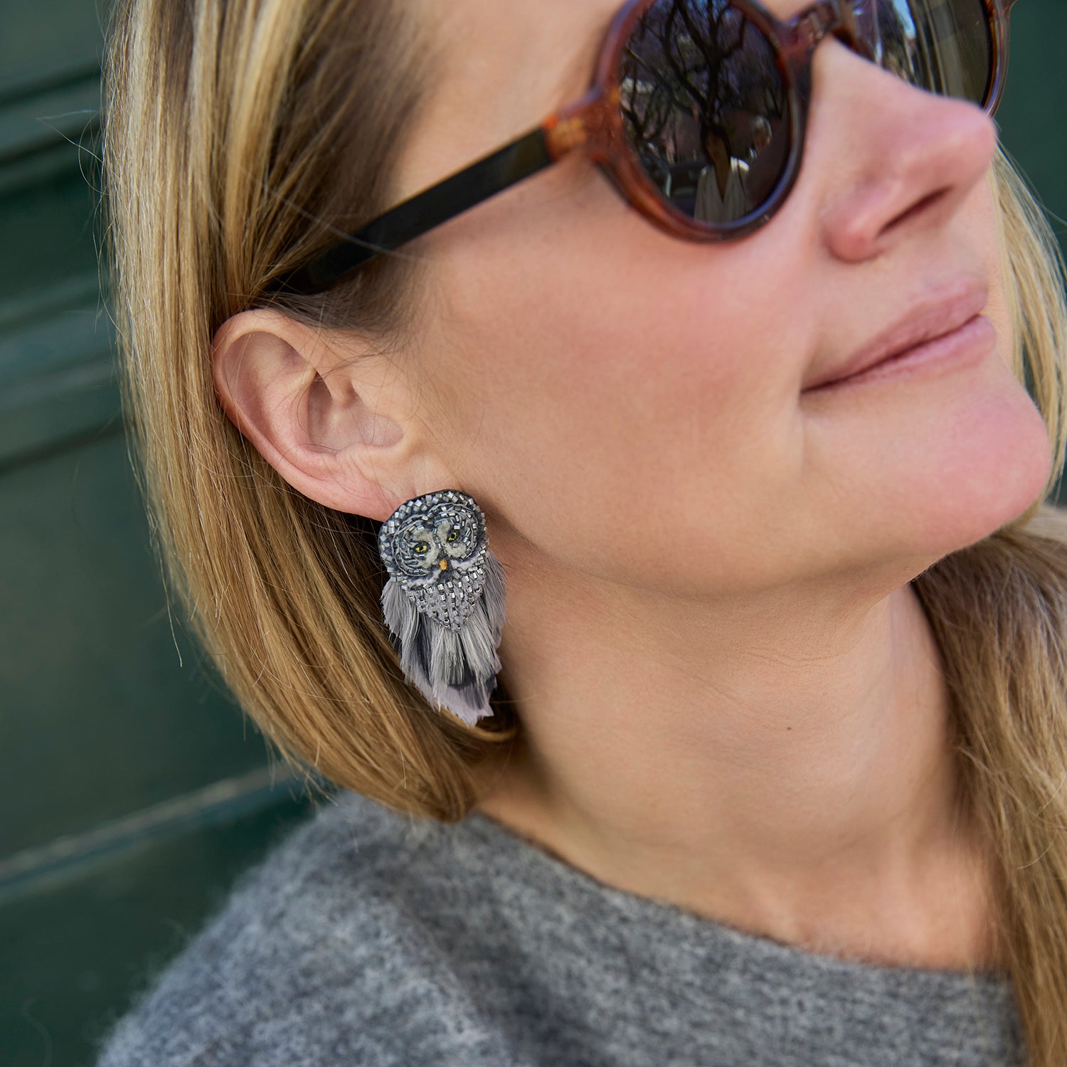 A close-up lifestyle shot of a woman wearing a silver owl-shaped dangle earring with white feathers, shown on her ear. She is wearing round sunglasses and a gray knit top.
