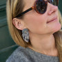A close-up lifestyle shot of a woman wearing a silver owl-shaped dangle earring with white feathers, shown on her ear. She is wearing round sunglasses and a gray knit top.