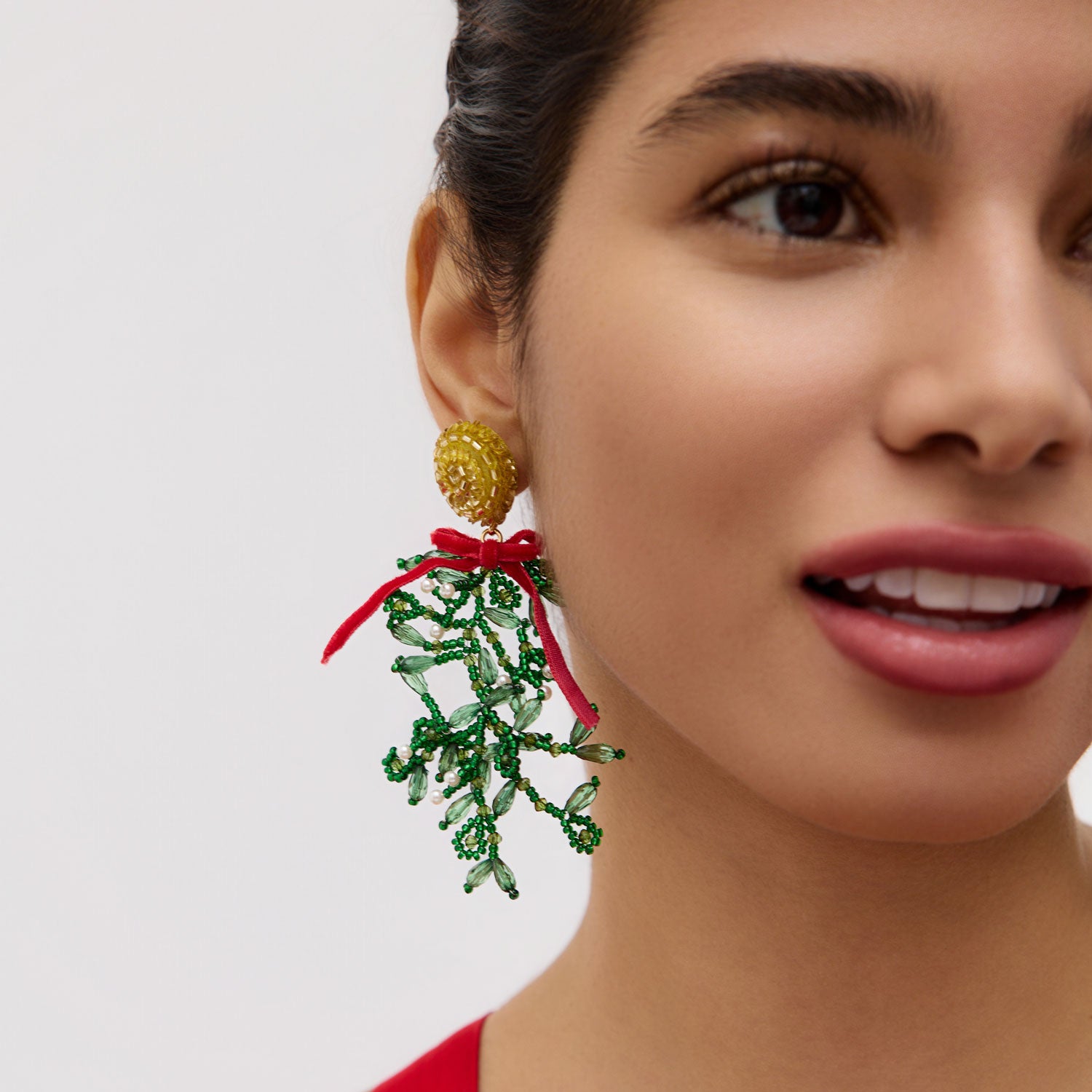 Close-up of a woman wearing a festive mistletoe earring with green leaves and a red bow on a plain background