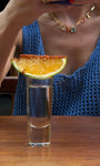 Glass of clear liquid with a lime and orange wedge garnish on a wooden surface, with a person in a blue top and gemstone chain necklace in the background.