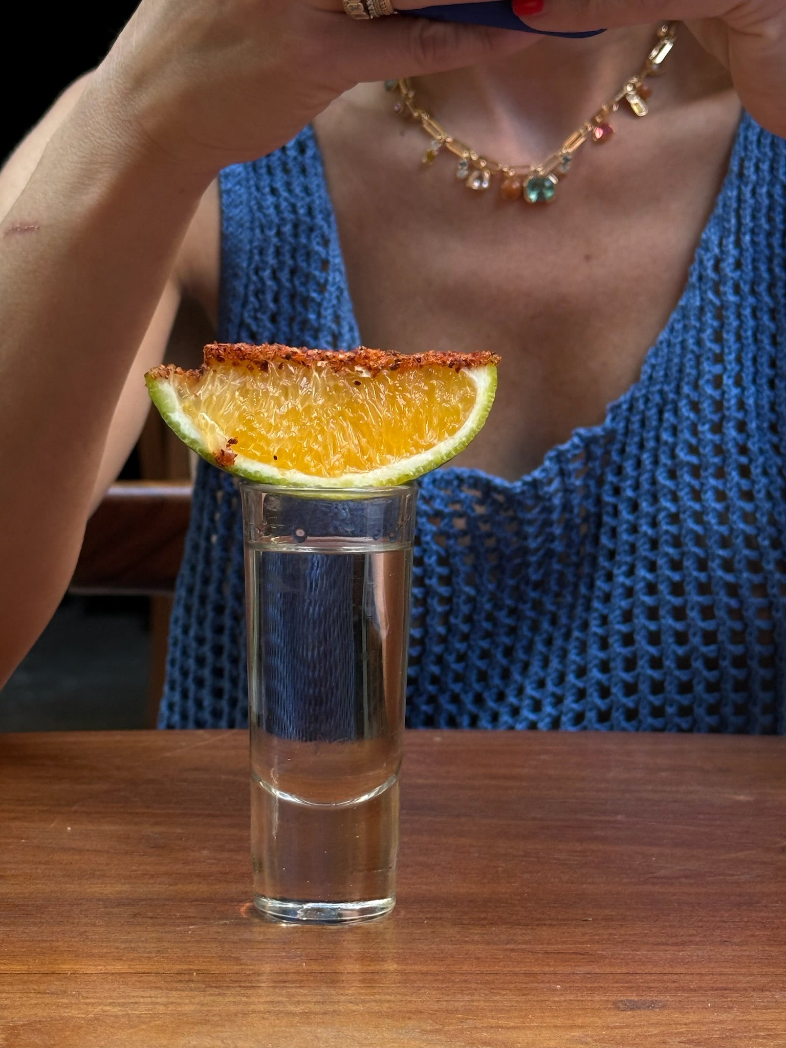 Glass of clear liquid with a lime and orange wedge garnish on a wooden surface, with a person in a blue top and gemstone chain necklace in the background.