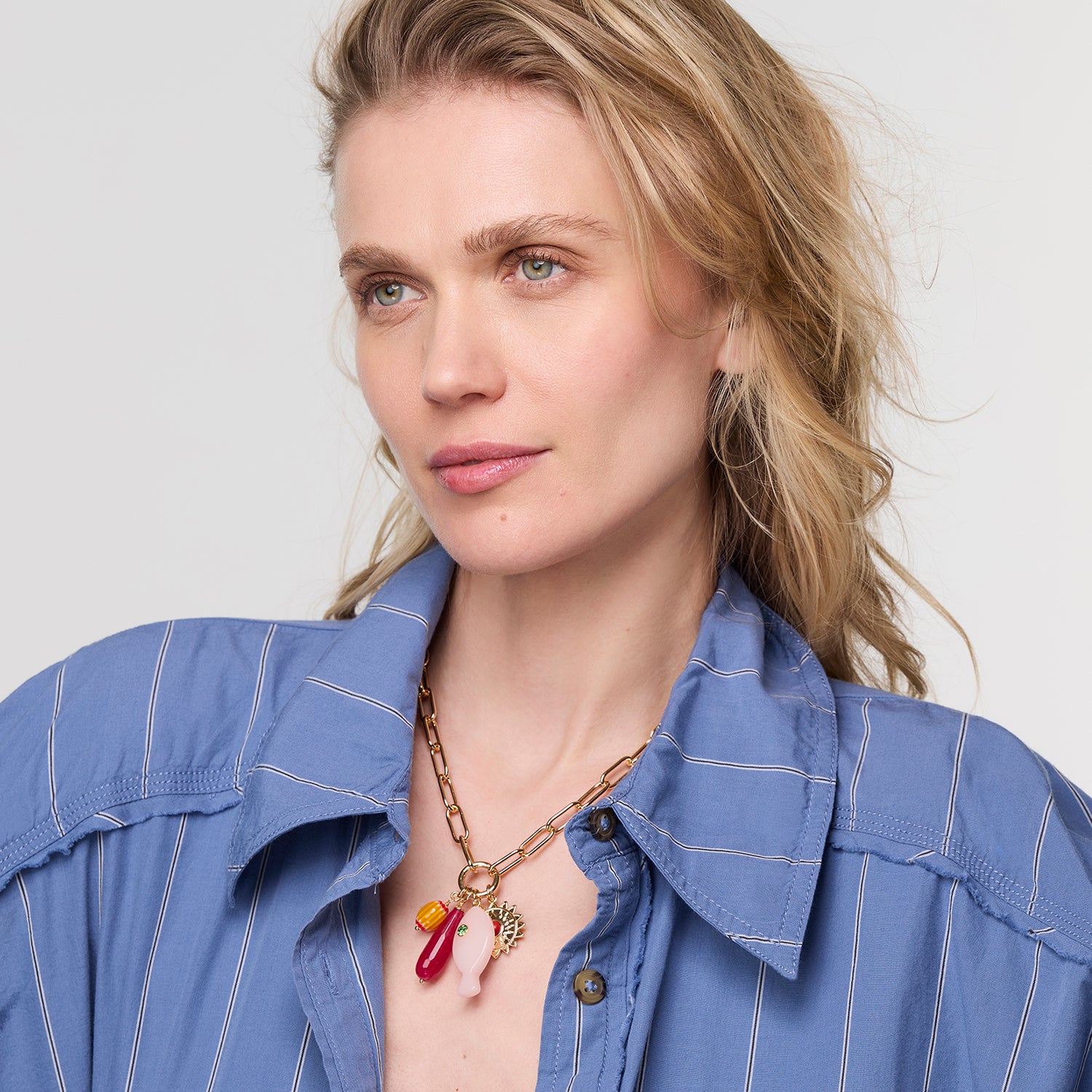 Woman wearing a blue shirt with a necklace featuring a red pendant against a plain background