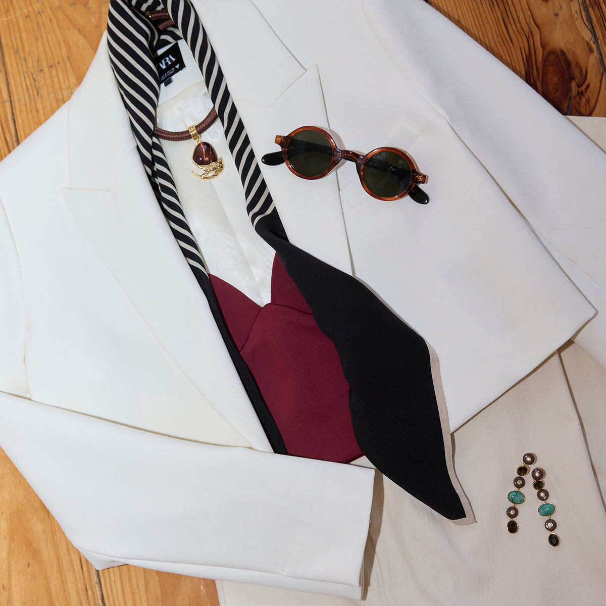 A styled flat-lay of a white blazer on a wooden floor, featuring a black and white striped necktie, a burgundy and black wingtip tie, tortoiseshell sunglasses, and a gold heart pendant necklace.