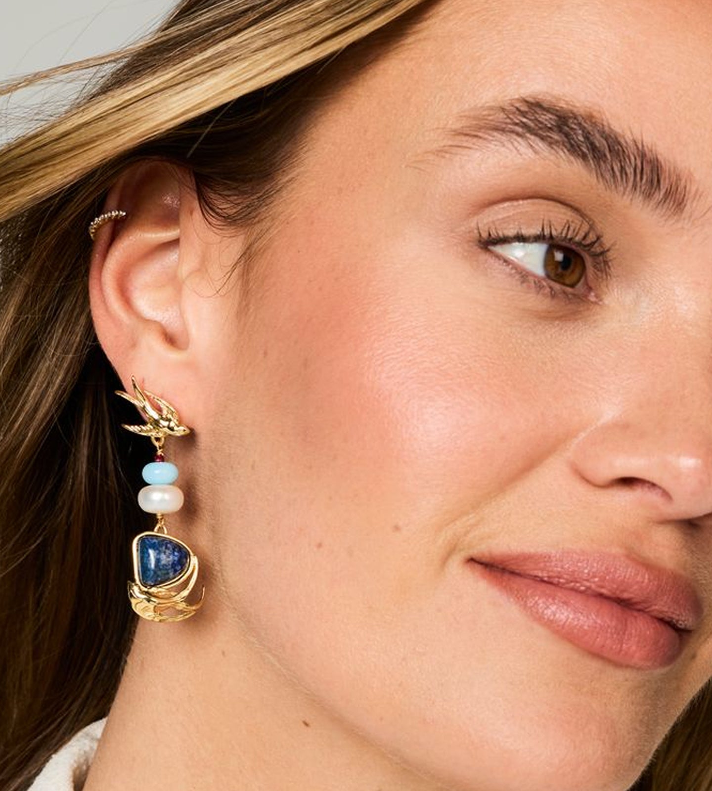 A close-up of a woman's ear wearing a gold dangle earring with a blue stone, light blue beads, and white pearl-like beads.