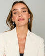 A woman with light brown hair wearing a white blazer and dangling blue and gold earrings, photographed against a plain light gray background.
