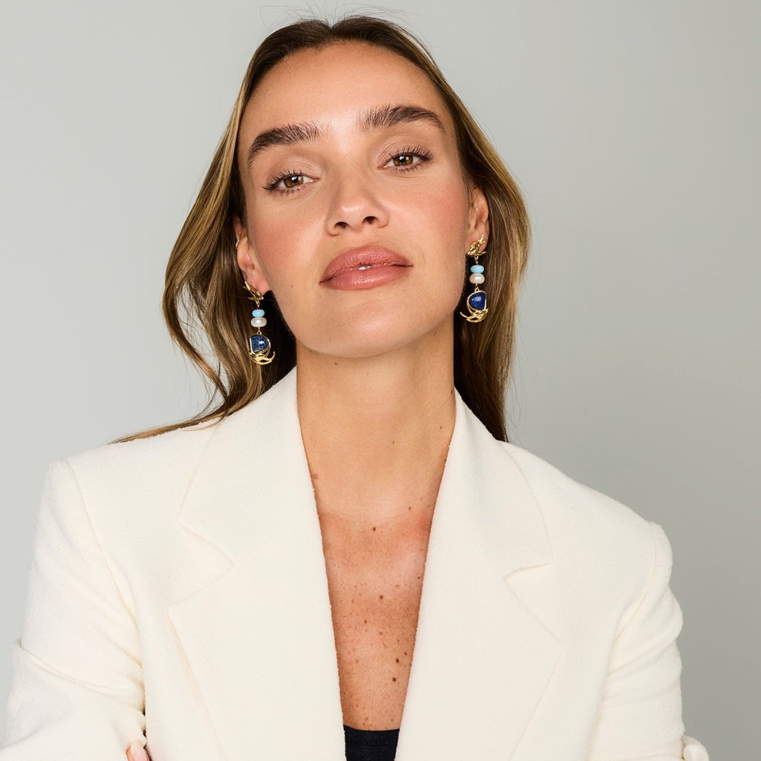 A woman with light brown hair wearing a white blazer and dangling blue and gold earrings, photographed against a plain light gray background.