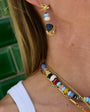Close-up of a person wearing a gold dangle earring with a dark blue stone and white beads, along with a beaded necklace and chain necklace against a green tiled background.
