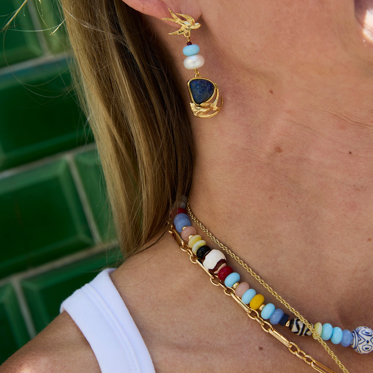 Close-up of a person wearing a gold dangle earring with a dark blue stone and white beads, along with a beaded necklace and chain necklace against a green tiled background.