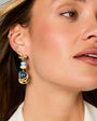 A close-up of a woman wearing a gold dangle earring with a blue heart-shaped stone, white pearl beads, and gold leaf details.