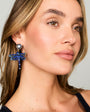A close-up of a woman wearing a dangling dragonfly earring made of blue rhinestones with a gold-toned setting and heart-shaped stud.
