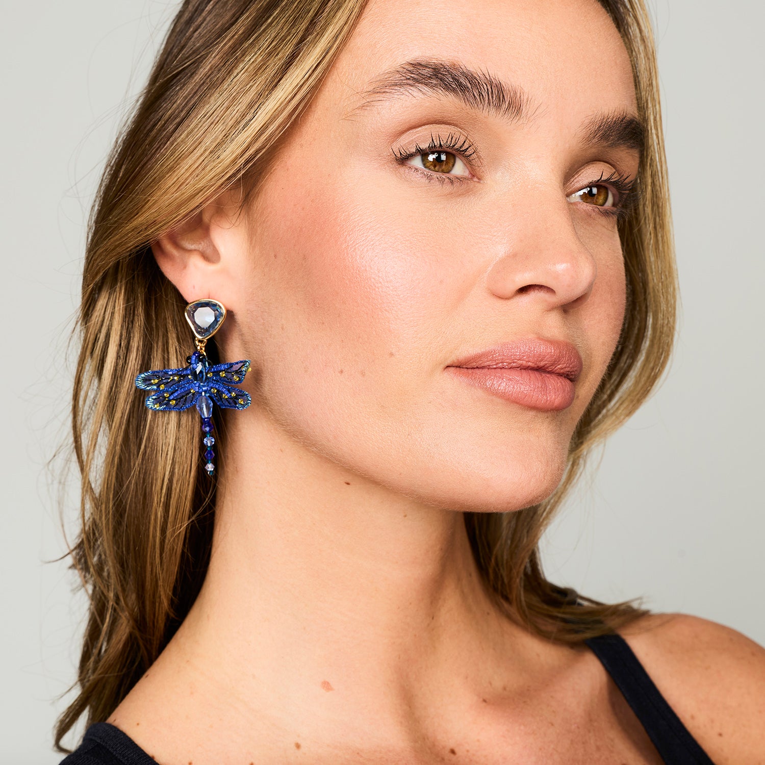 A close-up of a woman wearing a dangling dragonfly earring made of blue rhinestones with a gold-toned setting and heart-shaped stud.